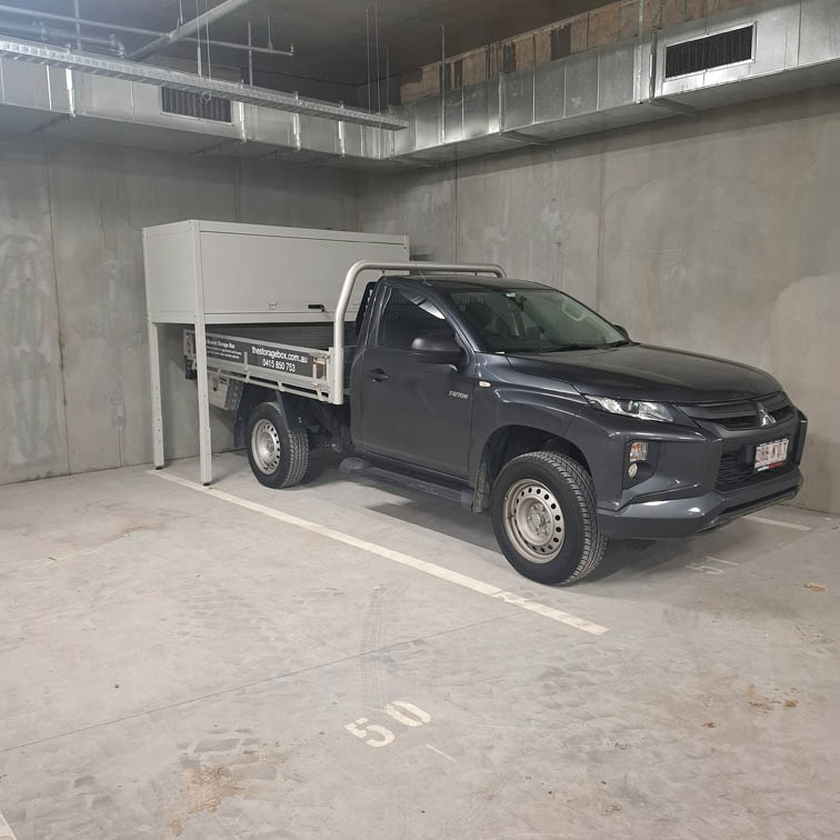 A dark grey utility truck is parked in an indoor concrete parking garage, occupying space number 50. The truck is parked under a metal storage box.