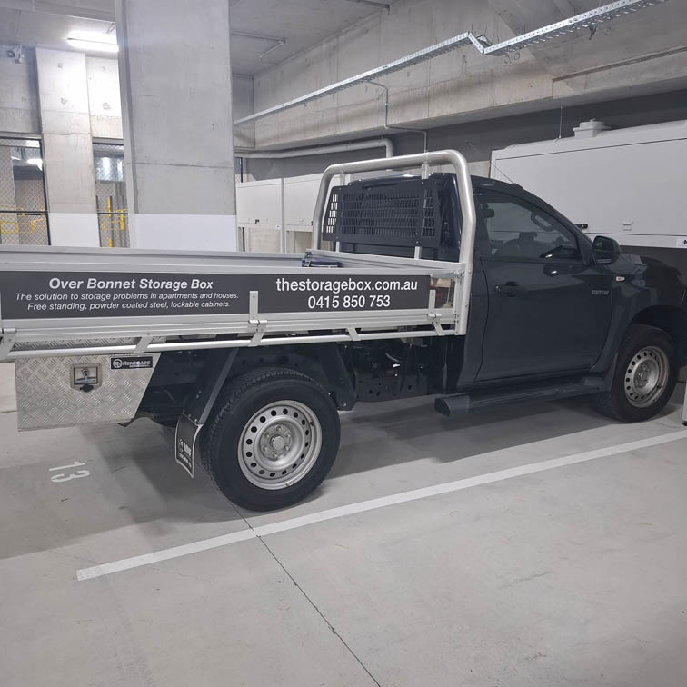 A black utility truck with advertising for Over Bonnet Storage Box on its side, parked in an indoor parking garage. Text includes a website and phone number for thestoragebox.com.au.