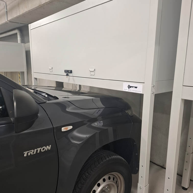 A black Triton vehicle is parked in a garage under a large, white, elevated metal storage cabinet with a closed front panel. The cabinet is mounted on legs above the car’s hood.