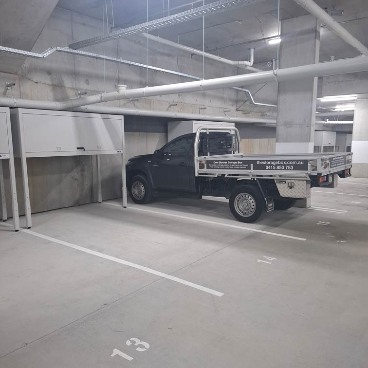 A black flatbed utility truck is parked in an indoor parking garage, occupying two spaces labeled 13 and 14 with over bonnet storage boxes. Overhead, pipes and lighting are visible along the concrete ceiling and walls.