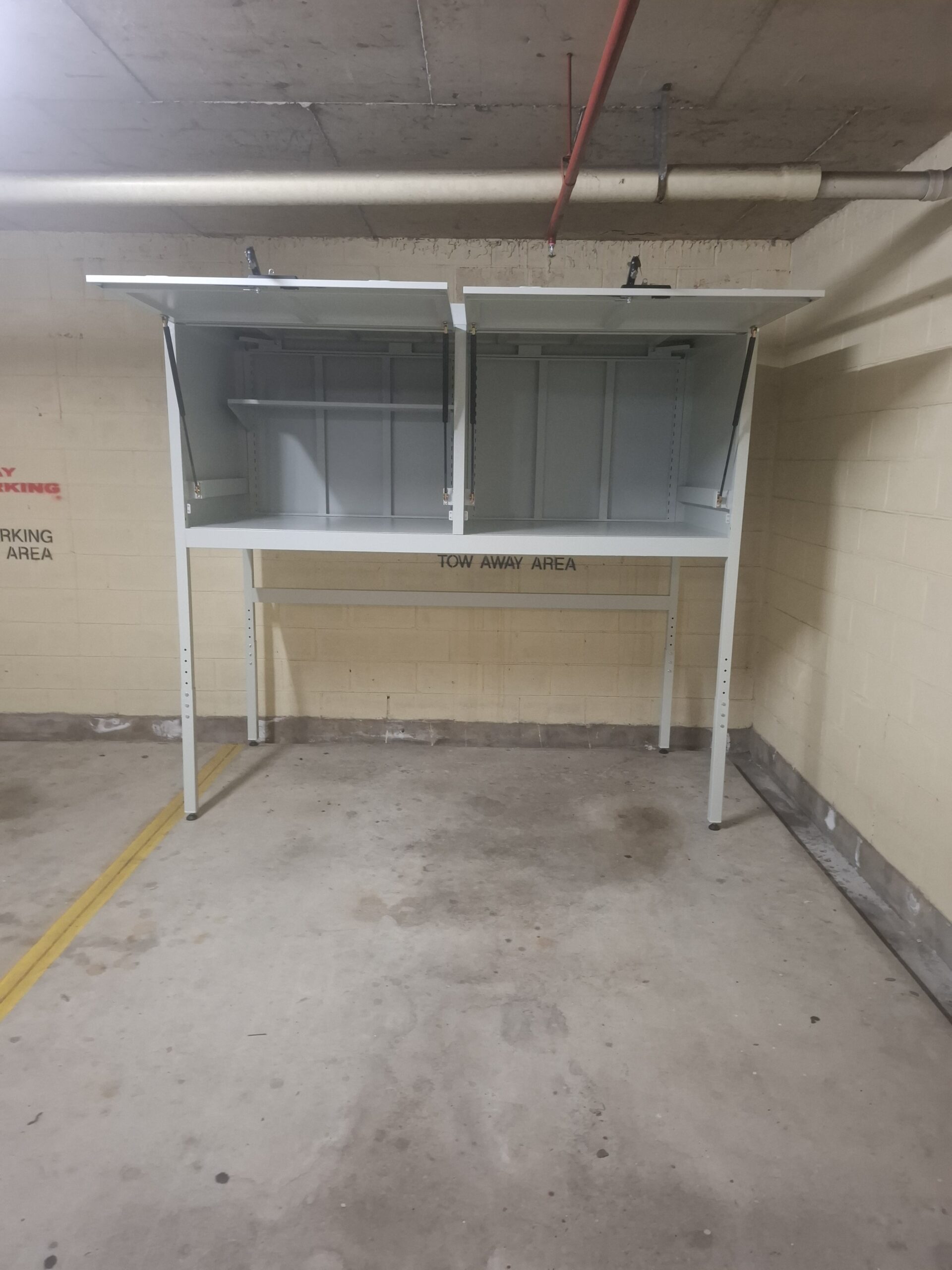 A 2 Door Over Bonnet Storage Cabinet with both doors open stands on tall legs in a concrete parking garage, featuring TOW AWAY AREA signage and yellow parking lines.