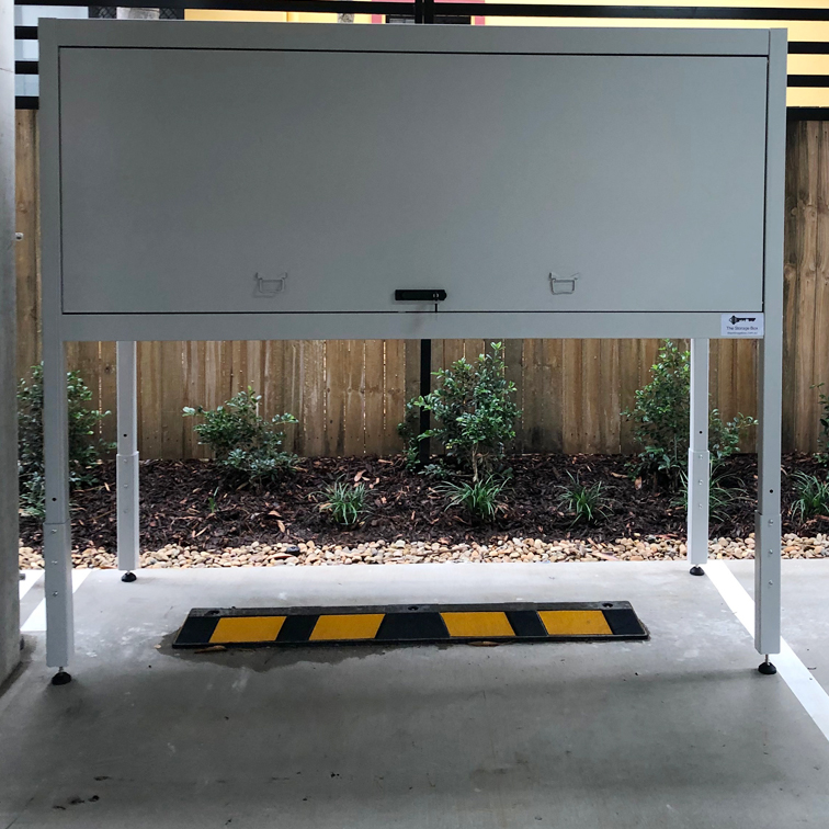 A large, gray metal storage cabinet on legs stands on a concrete surface, with a black and yellow wheel stop in front, small plants and a wooden fence in the background—offering an organized storage solution for outdoor spaces.